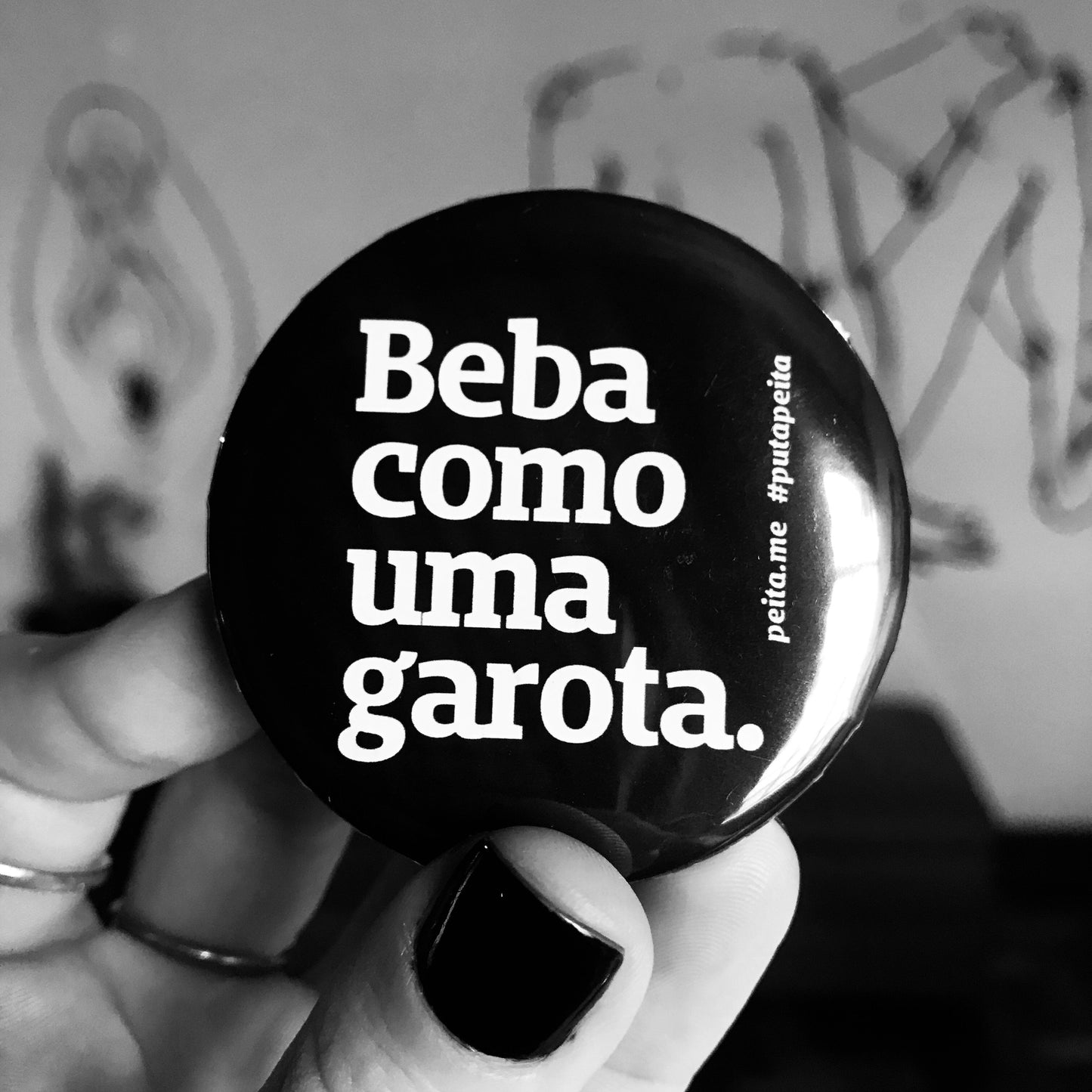 Foto preta e branca de um abridor preto, redondo, com a frase beba como uma garota em branco sendo segurado por uma mão branca. 