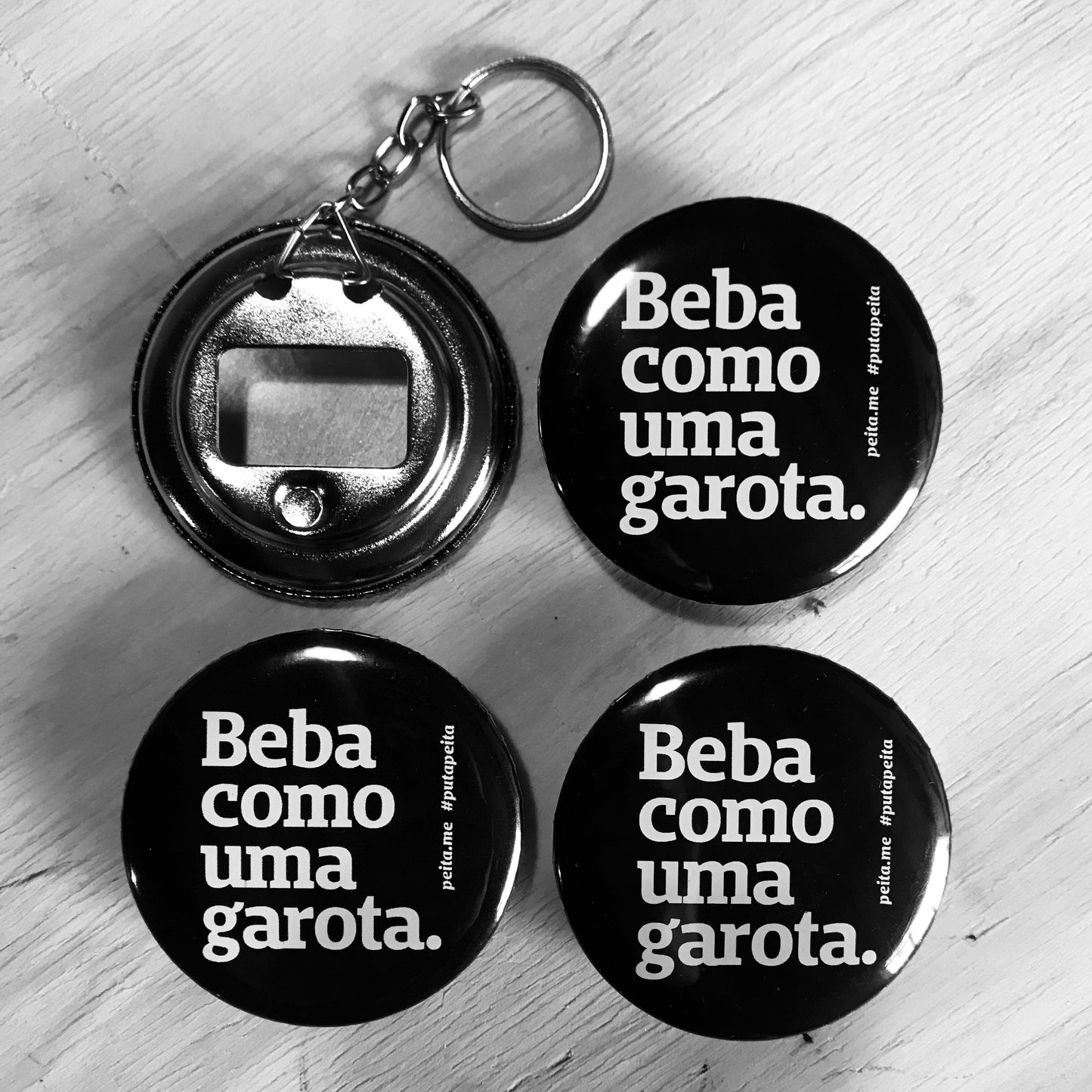 Foto preto e branca de 4 abridores de garrafa chaveiros redondos, pretos, com a frase lute como uma garota em branco. Um deles está virado, mostrando o encaixe do abridor.