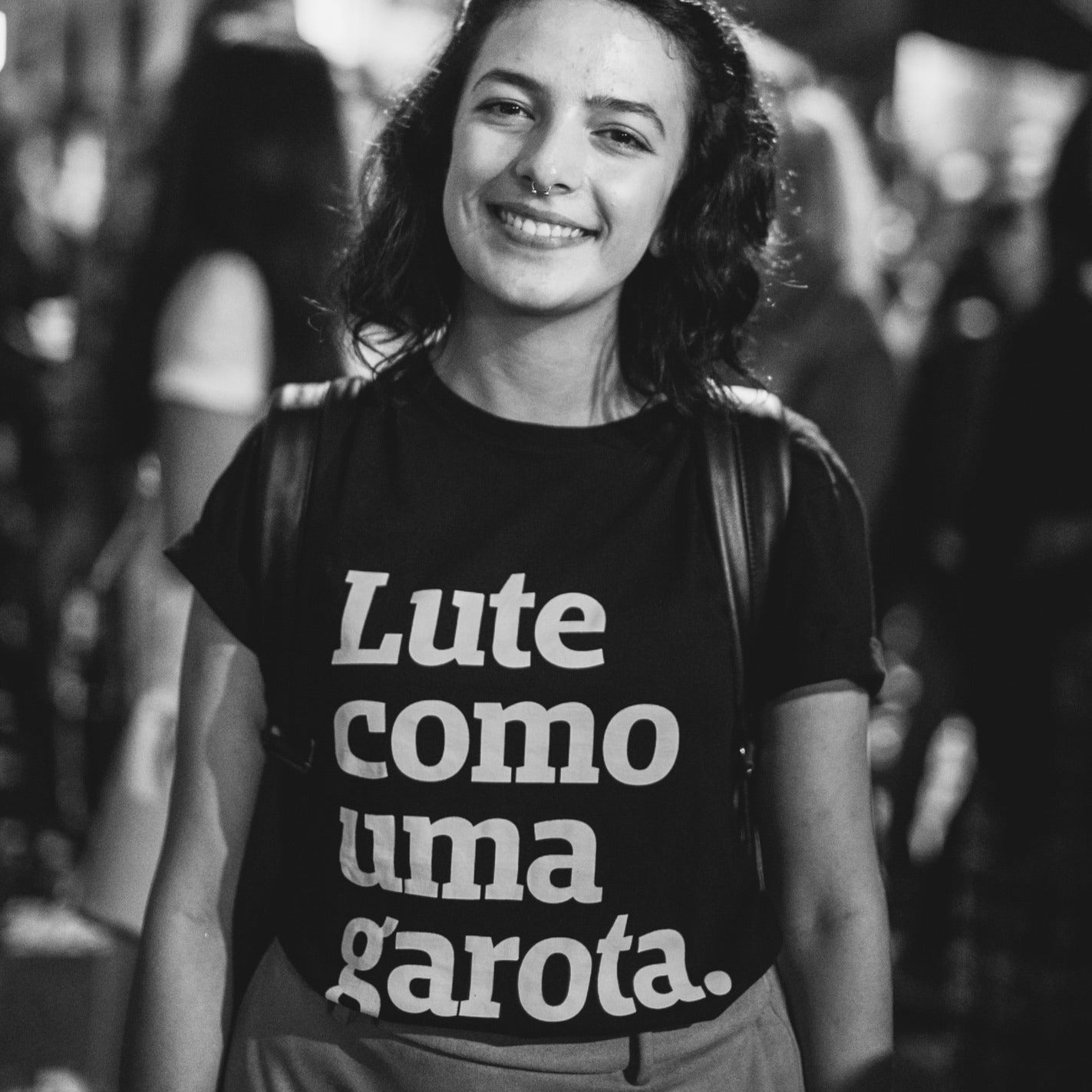 Foto preto e branco de mulher branca de cabelos escuros channel soltos, veste camiseta preta com frase Lute como uma garota em branco, e mochila nas costas. Ela sorri. Fundo desfocado