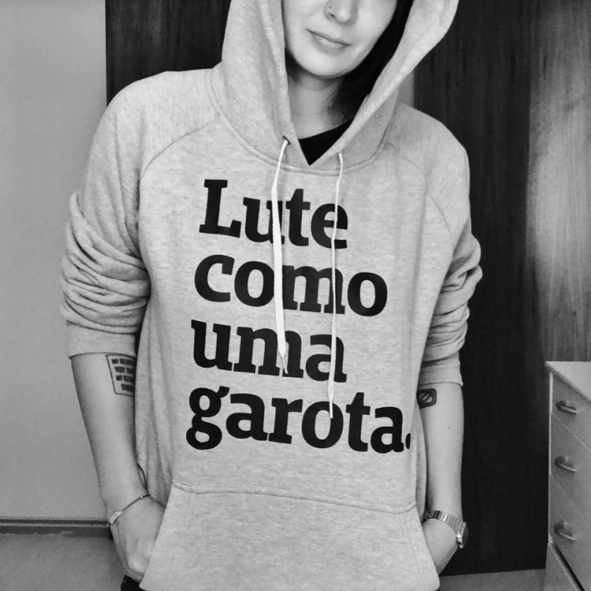 Foto preta e branca do tronco de uma mulher branca, com tatuagem no braço, vestida com moletom cinza escrito lute como uma garota em preto. Está com o capuz na cabeça.