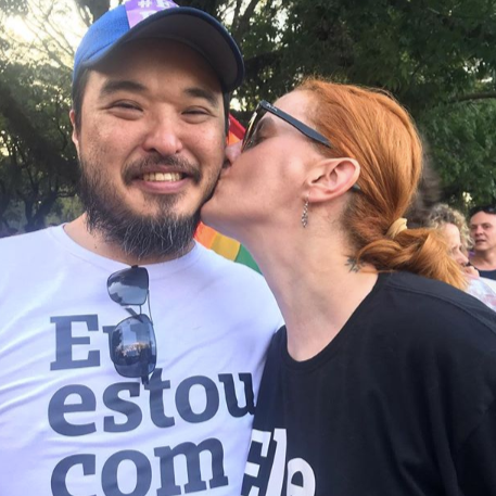 Foto colorida de uma mulher branca com cabelos ruivos amarrado e um homem oriental, com barba escura com fio grisalhos. Ela usa camiseta preta escrita ele não em branco e ele usa camiseta branca escrita eu estou com ela em preto. Ela está beijando a bochecha dele.