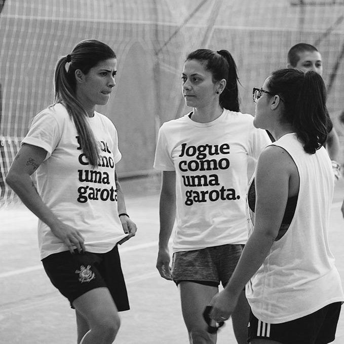 Foto preta e branca de 3 mulheres brancas. A do meio tem cabelos escuros, lisos, médios, presos em rabo. A da esquerda tem cabelos lisos, escuros nas raízes e loiros nas pontas.  A da direita tem cabelo liso, escuro, preso em rabo, usa óculos. Ambas vestem peita branca com a frase jogue como uma garota em preto. 