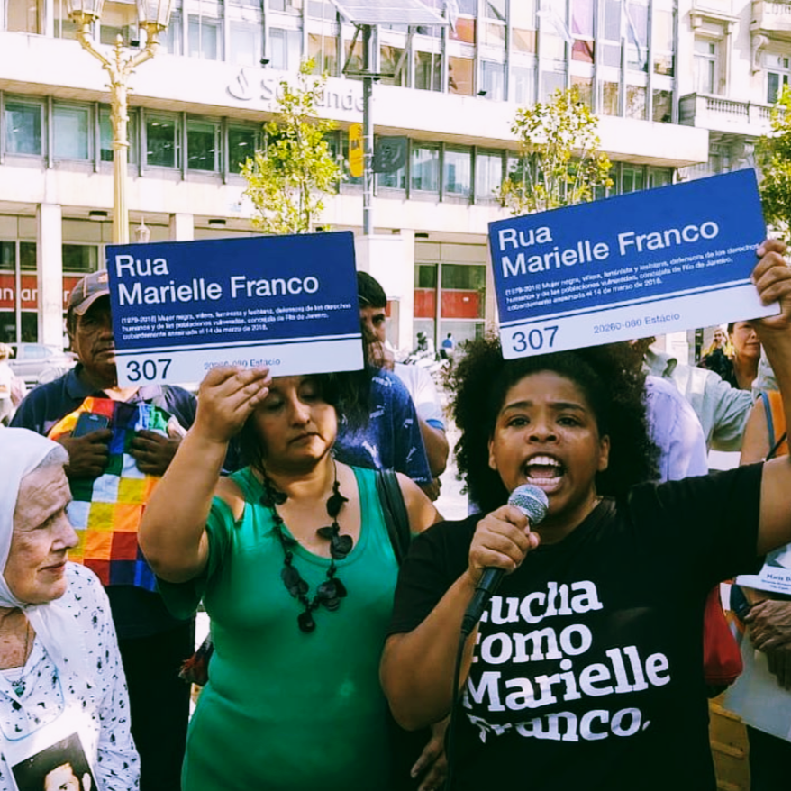 Foto colorida de uma mulher negra, cabelo crespo, preto, médio, com arco, está vestida com camiseta preta escrita lucha como Marielle Franco em branco, com mão esquerda segura um microfone próximo a boca. A sua esquerda,uma mulher branca, cabelo castanho, liso, chanel curto, com olhos fechados e blusa verde. Ambas estão com placas azuis escritas Rua Marielle Franco Número 307 em branco.