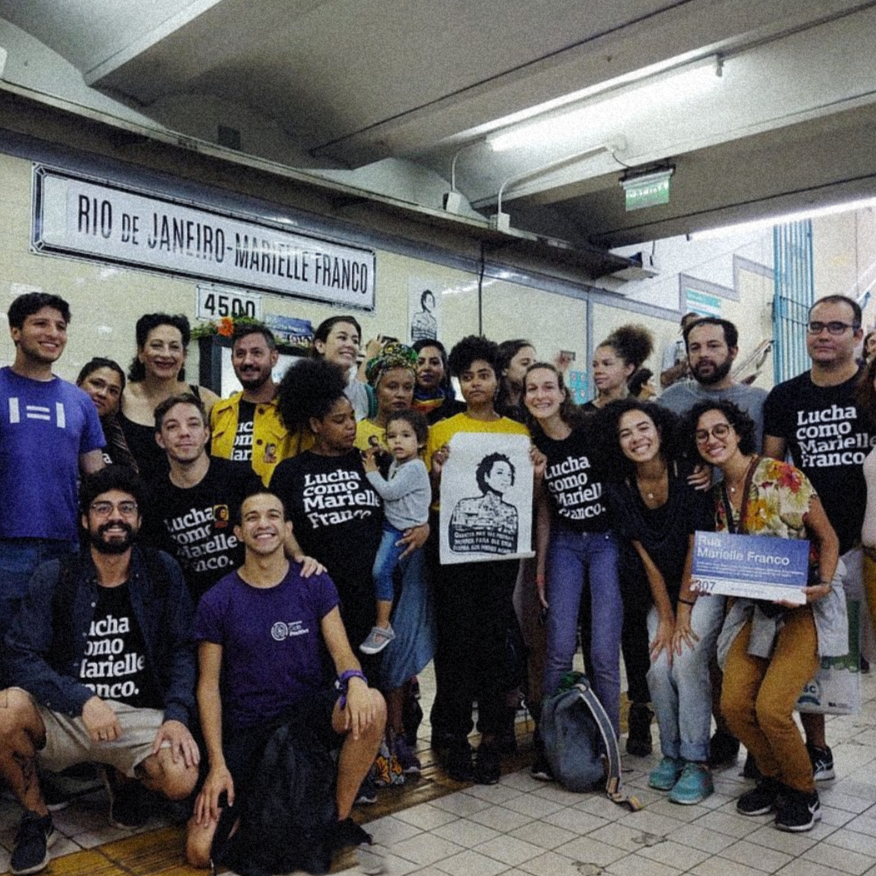Foto colorida de várias pessoas, mulheres brancas e negras, homens brancos e negros. Nela, cinco pessoas vestem peita preta com a frase lucha como Marielle Franco em branco. Uma das mulheres segura uma placa de rua com o nome de Marielle Franco e as pessoas estão paradas na estação de metrô Rio de Janeiro - Marielle Franco, em Buenos Aires.