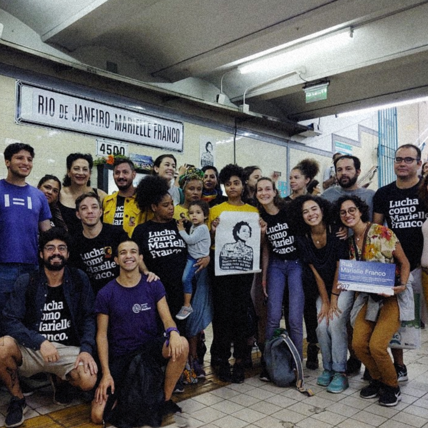  Foto colorida de várias pessoas, mulheres brancas e negras, homens brancos e negros. Nela, cinco pessoas vestem peita preta com a frase lucha como Marielle Franco em branco. Uma das mulheres segura uma placa de rua com o nome de Marielle Franco e as pessoas estão paradas na estação de metrô Rio de Janeiro - Marielle Franco, em Buenos Aires.