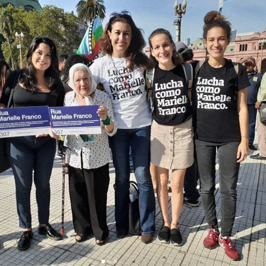 Foto colorida de 5 mulheres brancas, da direita para esquerda:mulher branca, cabelo loiro, chanel; mulher, cabelos castanhos, crespos, presos em coque; mulher branca, cabelos castanhos, lisos, longos; no meio, mulher branca, cabelos castanhos, lisos, longos, vestidas camiseta escrita Lucha como Marielle Franco em branco. A penúltima, Nora Cortiñas, lenço branco na cabeça e última tem cabelos castanhos, lisos, longos e soltos. Ambas seguram placas azuis de rua escritas Rua Marielle Franco, n° 307 em branco.