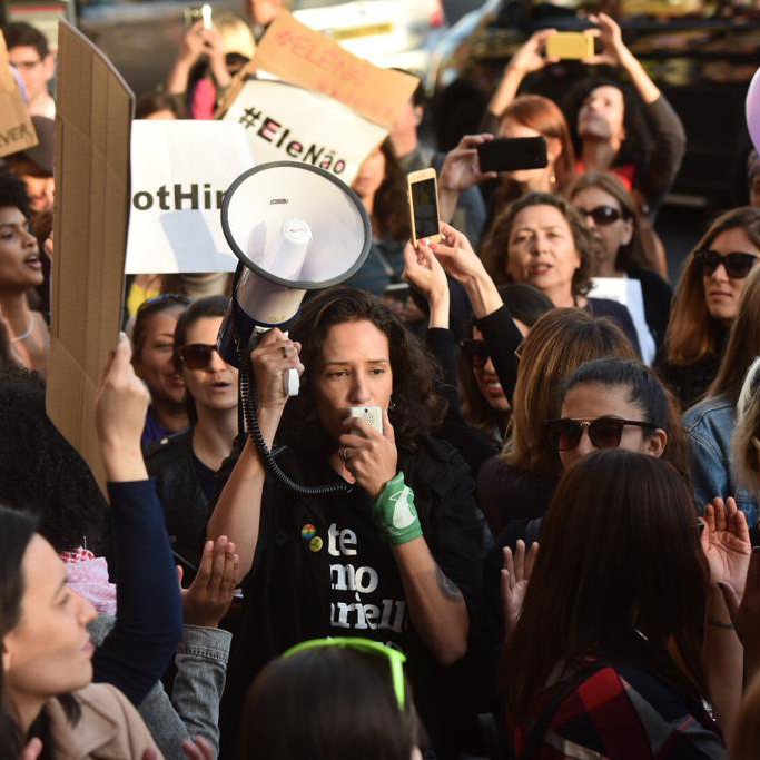 Foto colorida de uma mulher branca, com cabelos castanhos, cacheados, chanel, vestida com camiseta escrito lute como Marielle Franco em branco, com uma jaqueta preta  por cima. Está segurando um megafone na mão direita e fala com  outras pessoas no meio de uma multidão.