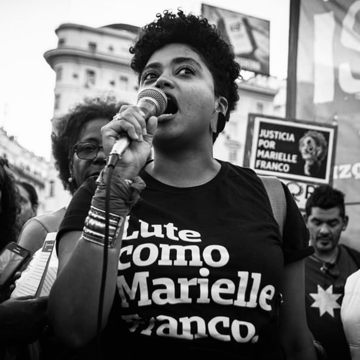 Foto branca e preta de uma mulher negra, cabelos crespos, pretos, curtos, está vestida com camiseta preta escrito lute como Marielle Franco branco.  No pulso esquerdo tem uma pulseira e um lenço amarrado,  na mão direita segura o microfone.