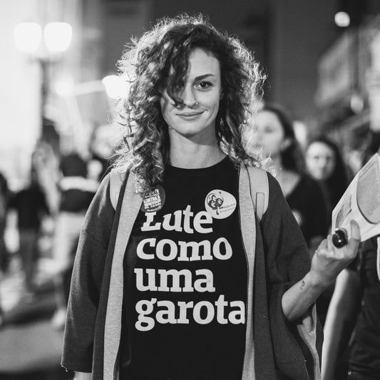 Foto preta e branca de uma mulher branca, cabelos castanhos, cacheados, médios, vestida com camiseta preta escrita lute como uma garota em branco, usa um casaco por cima. Na mão direita segura papéis, sorri para a foto.