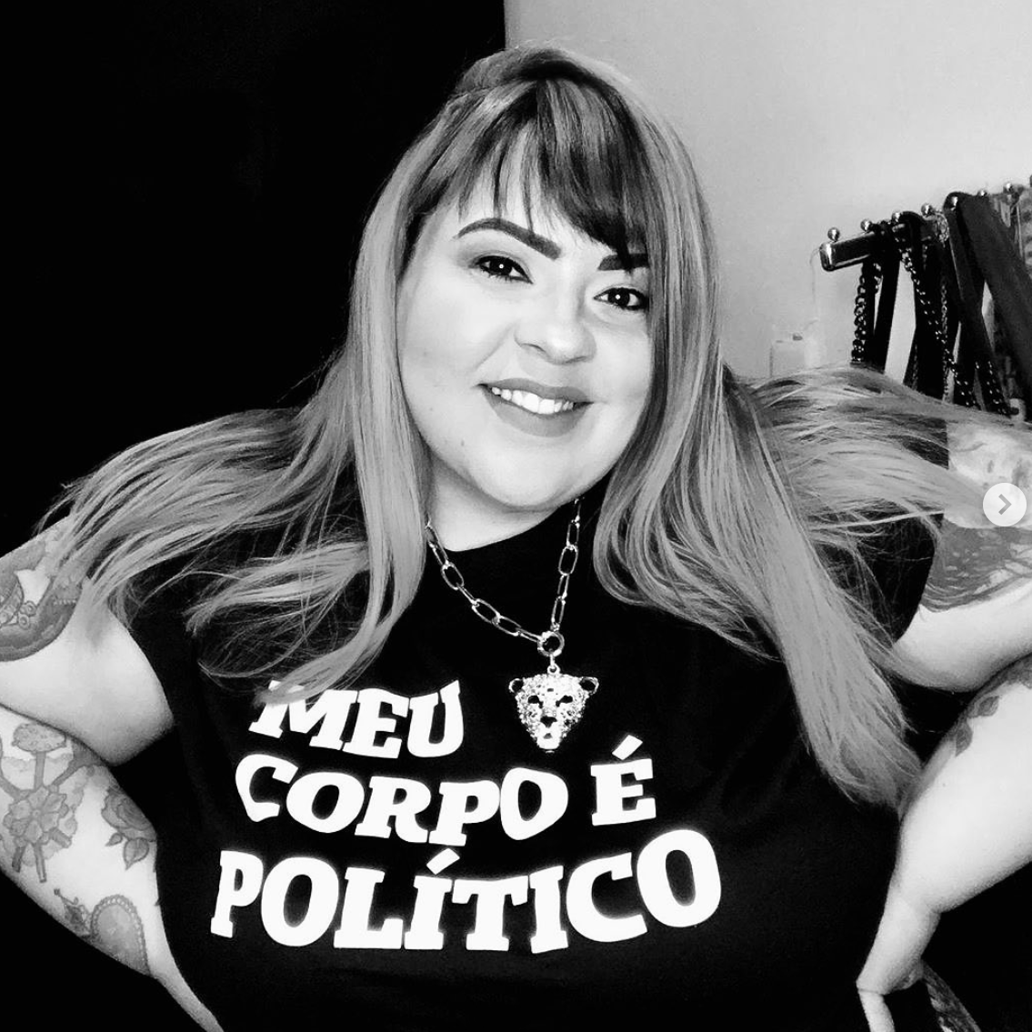 Foto preta e branca de uma mulher branca, com cabelos lisos, castanhos com mechas loiras, franja, longos, com braços tatuados, está vestida com camiseta preta escrita meu corpo é político em branco. Suas mãos estão na cintura e sorri para foto.