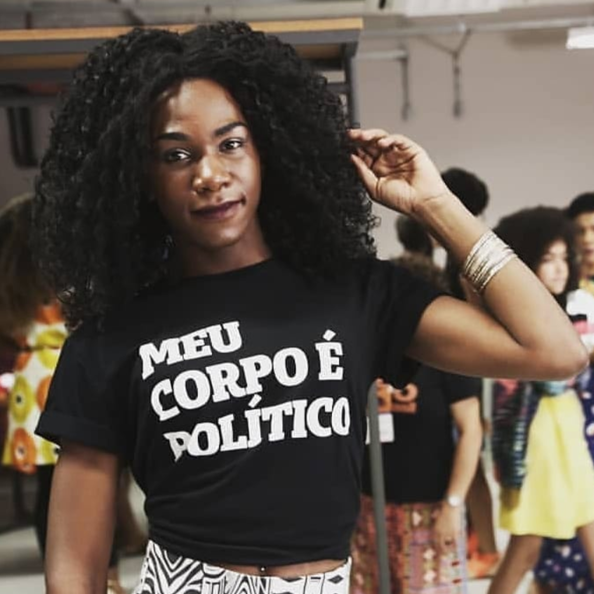 Foto colorida de uma mulher negra, cabelos cacheados, pretos, médios, soltos, usa pulseira no pulso esquerdo, está vestida com camiseta preta escrita meu corpo é político em branco. Segura  os cabelos com a ponta dos dedos.