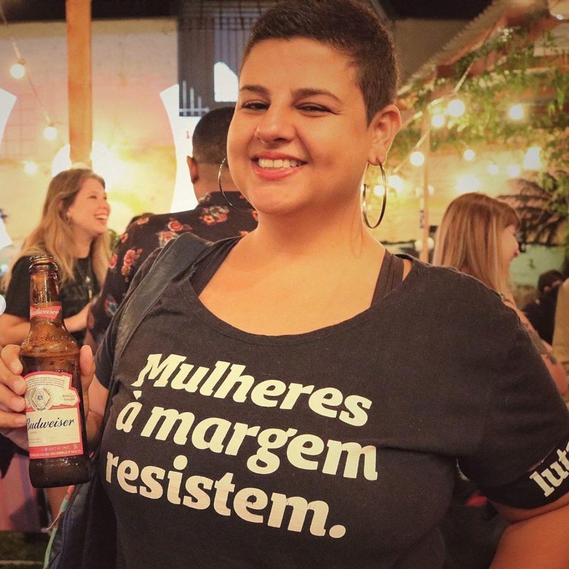 Foto colorida de uma mulher branca, cabelos castanhos, lisos, curtos, vestida com camiseta preta escrita mulheres à margem resistem em branco. Ela segura uma cerveja na mão esquerda e sorri para a foto.