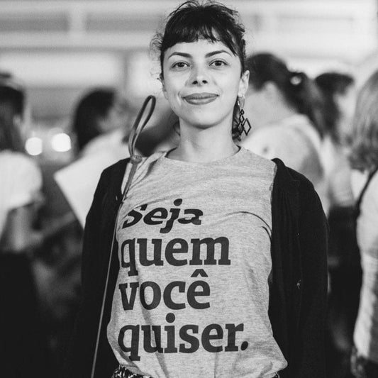 Foto preta e branca de uma mulher branca, cabelos presos, castanhos com franja,vestida com camiseta cinza escrita seja quem você quiser em preto, com um casaquinho preto por cima. Está sorrindo na foto.