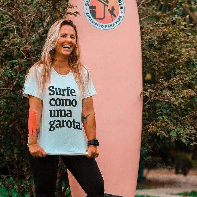 Foto colorida de uma mulher branca, cabelo loiro, liso, comprido, solto, tatuagens nos braços, relógio preto no pulso direito, vestida com camiseta branca escrita surfe como uma garota em preto. Ela puxa a barra para baixo, sorri na foto. Atrás tem uma prancha rosa.