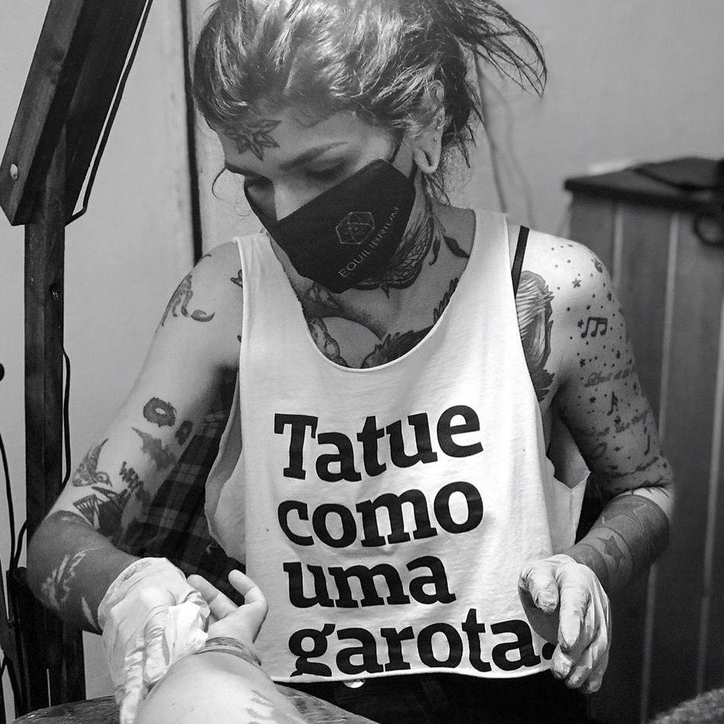 Foto preta e branca de uma mulher branca, cabelos pretos, lisos, presos, com tatuagem na testa, nos braços, peito e pescoço, usa máscara preta, está vestida com camiseta branca escrita tatue como uma garota em preto. Está higienizando um braço recém tatuado.