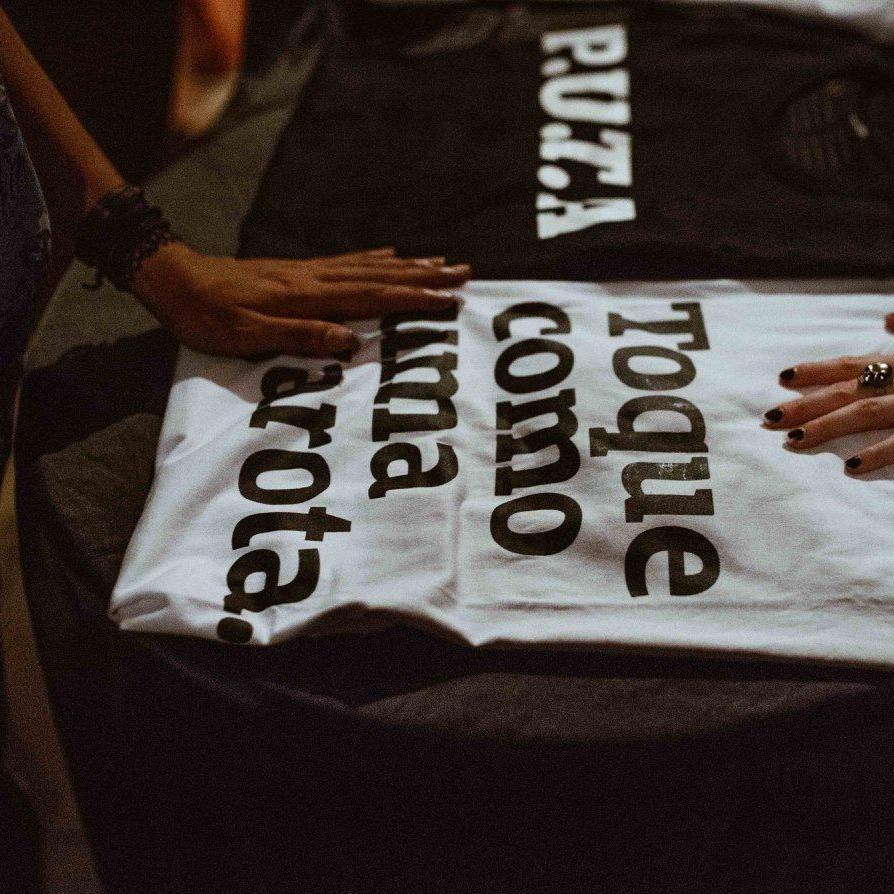 Foto colorida de 2 camisetas dobradas em cima de um barril, uma preta escrita P.U.T.A. em branco e outra branca escrita toque como uma garota em preto. Aparecem 2 mãos brancas tocando.