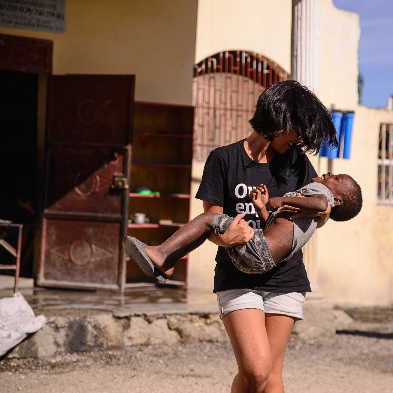 Foto colorida de uma mulher branca, cabelos castanhos, lisos, chanel, vestido com camiseta preta escrita você importa pra mim em branco. Ela está andando segurando uma criança negra no colo, ambas se olham e sorriem.