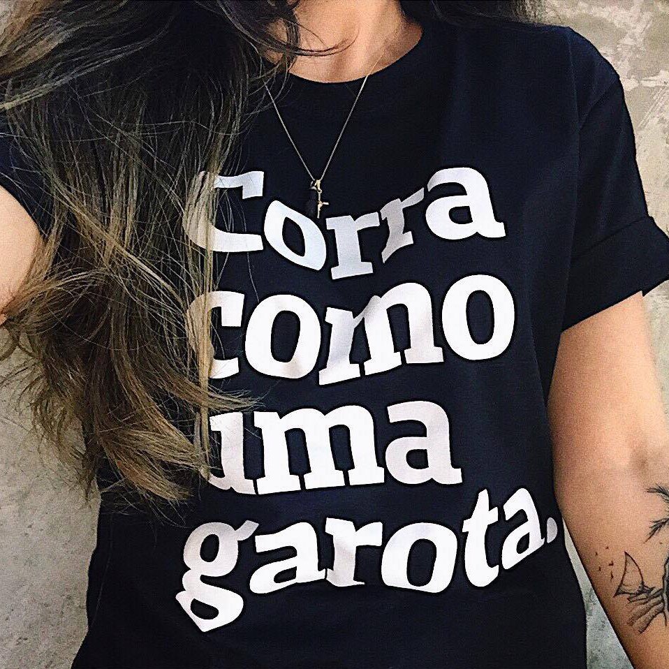 Foto colorida do tronco de uma mulher branca com cabelo castanho, liso e solto. Veste uma peita preta escrito corra como uma garota em branco.