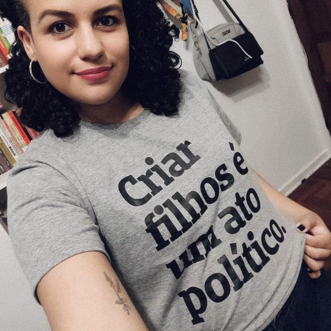 Foto colorida de uma mulher negra, com cabelo castanho, até os ombros, preso. Está com uma camiseta cinza com a frase criar filhos é um ato político em preto. Ela sorri pra foto.