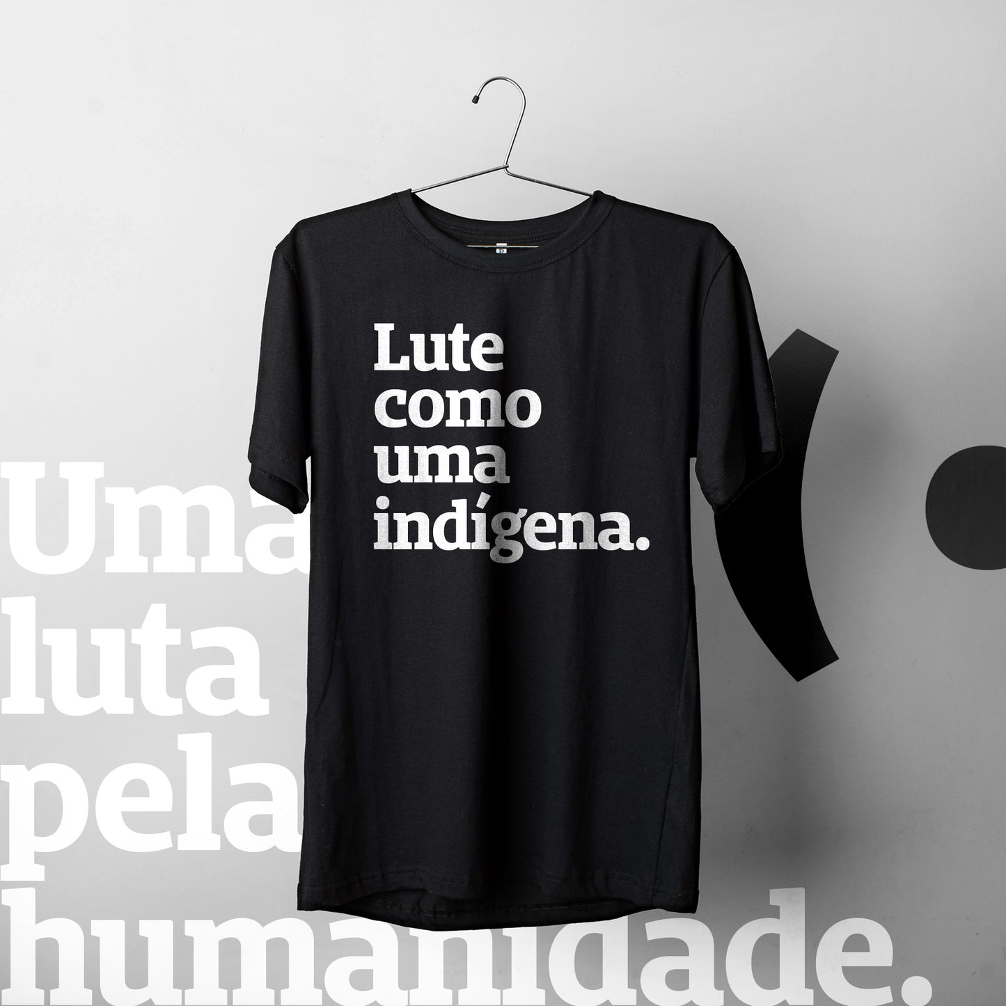 Foto colorida de camiseta preta com frase Lute como uma indígena em branco pendurada no cabide. Fundo mesclado de cinza claros com logo da peita em preto no lado direito e marca d'água com frase uma luta pela humanidade ao lado esquerdo