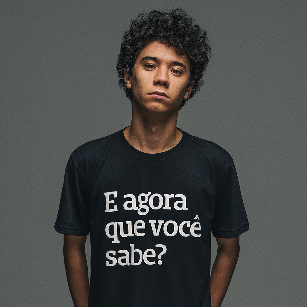 Foto colorida em estúdio de um homem negro, de cabelos escuros e encaracolados, veste camiseta preta com a frase E agora que você sabe? em branco. Olha sério pra foto