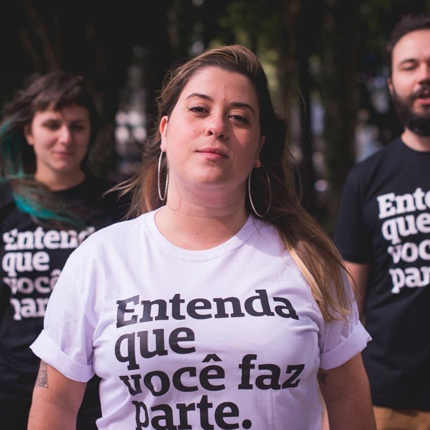 Foto colorida de 2 mulheres brancas, com cabelos lisos, compridos, castanho e loiro e 1 homem branco com cabelos lisos, curtos, castanhos e barba curta . A do meio está de camiseta branca escrito entenda que você faz parte em preto. , A mulher da sua esquerda e o homem,vestem  camisetas pretas com a frase entenda que você faz parte em branco Todas olham sério pra foto. 