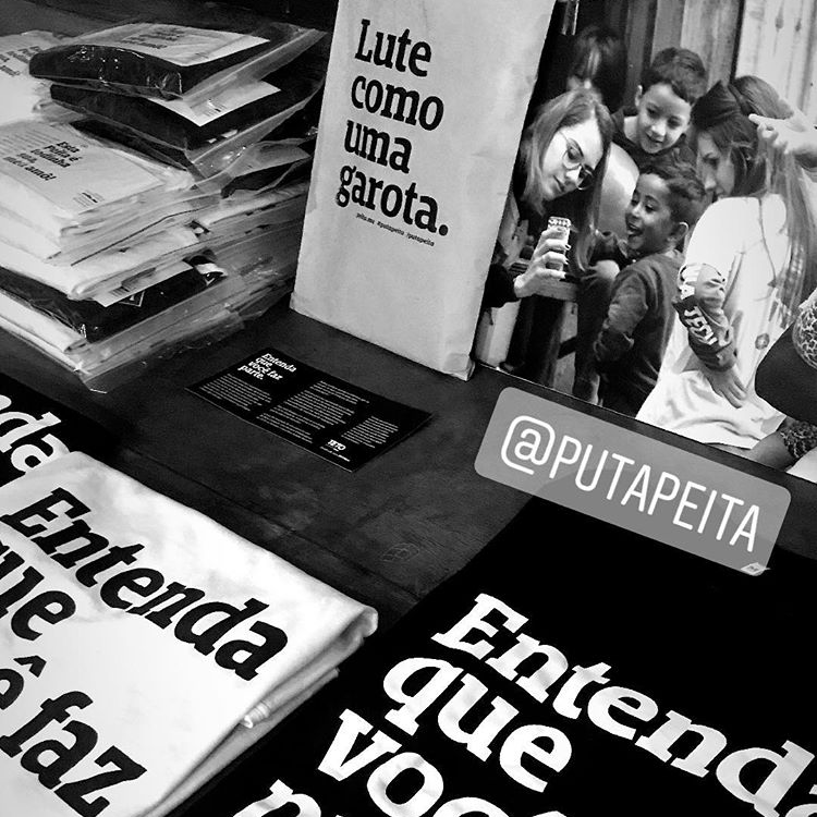 Foto preto e branca de camisetas nas cores branco com preto e preto com branco com a frase entenda que você faz parte expostas em uma mesa. Tem duas pilhas de camisetas embaladas e um cartaz branco escrito lute como uma garota