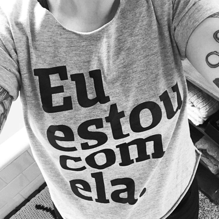 Foto preta e branca selfie de uma mulher branca com camiseta cinza escrita eu estou com ela em preto. Tem tatuagem no braço.