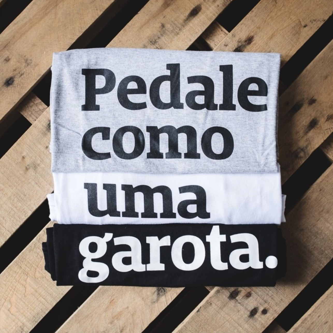 Foto colorida de três camiseta empilhadas formando a frase Pedale como uma garota, de cima para baixo tem Cinza com escrita preta, no meio branca com escrita preta e por ultimo preta com escrita branca. Fundo uma mesa em palete de madeira