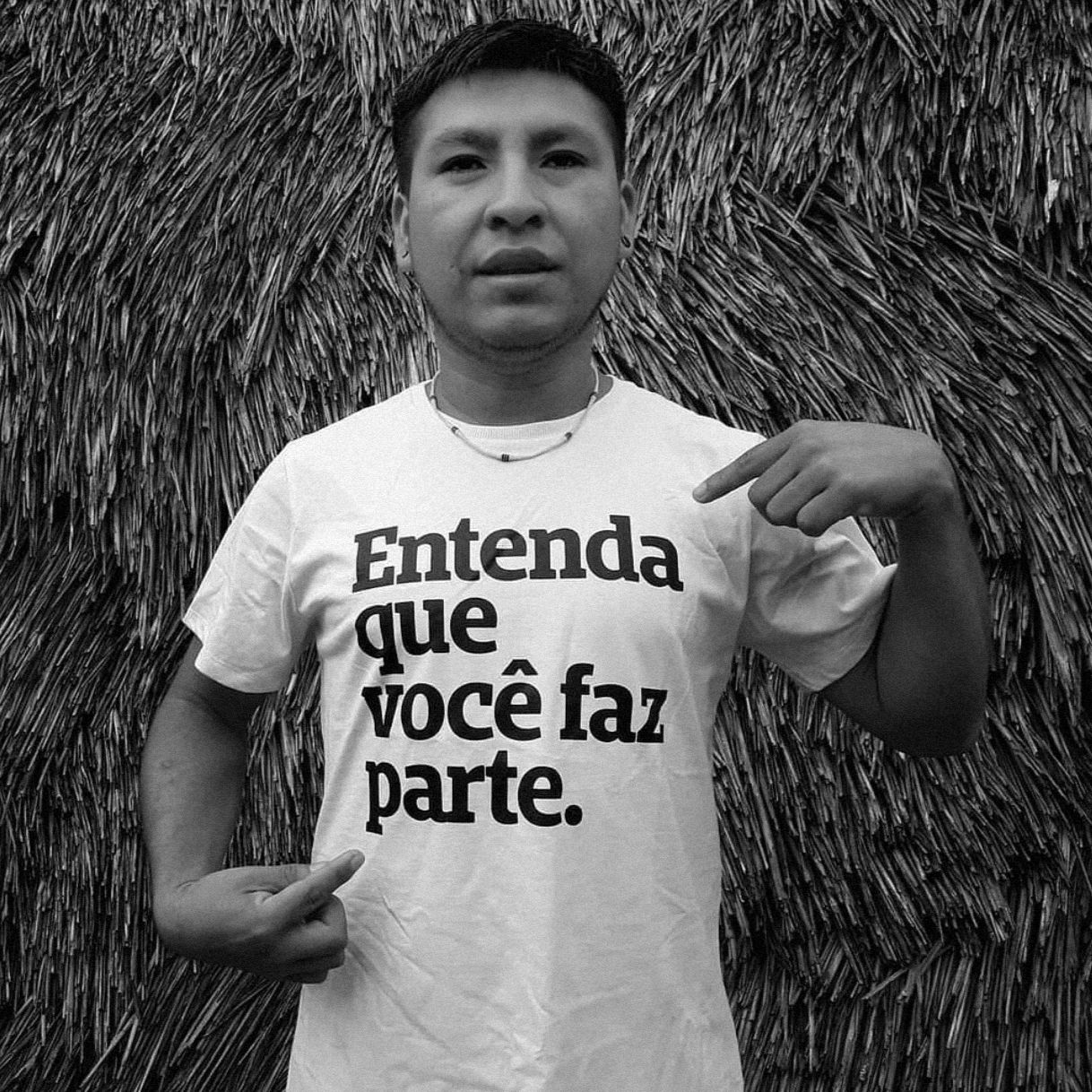 Foto preto e branco de homem indígena com camiseta branca escrito Entenda que você faz parte em preto. Usa colar e brinco, aponta com dedo indicador das duas mãos para a frase no corpo.