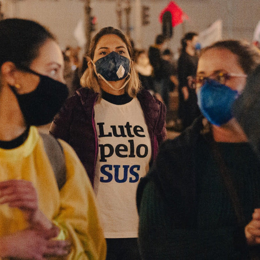 Foto colorida de mulher branca loira de cabelos lisos que veste camiseta branca com escrita Lute pelo sus em preto e azul, com jaqueta roxa por cima e mascara preta, Fundo desfocado