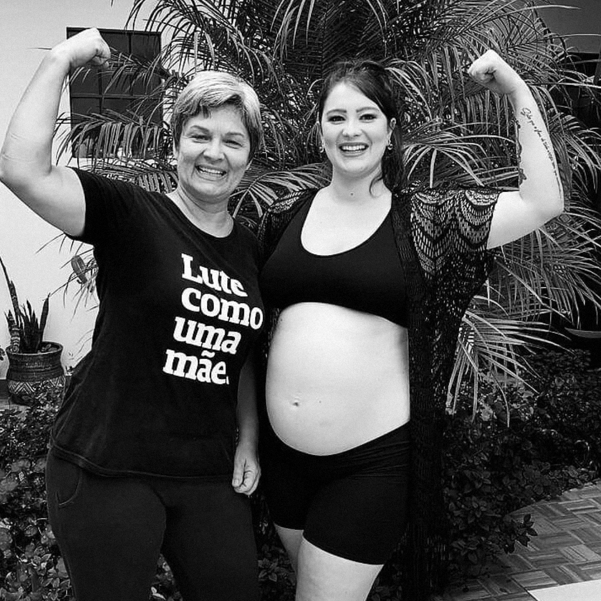 Foto preto e branco de duas mulheres abraçada0s. a da esquerda mulher branca de cabelos claros e curtos, veste camiseta preta com frase Lute como uma mãe em branco e ergue braço esquerdo em punho fechado, a outra mulher branca de cabelos escuros lisos, preso em rabo, veste top preto e está grávida, ergue mão direita fazendo punho fechado, ambas sorri. Fundo quintal com plantas.