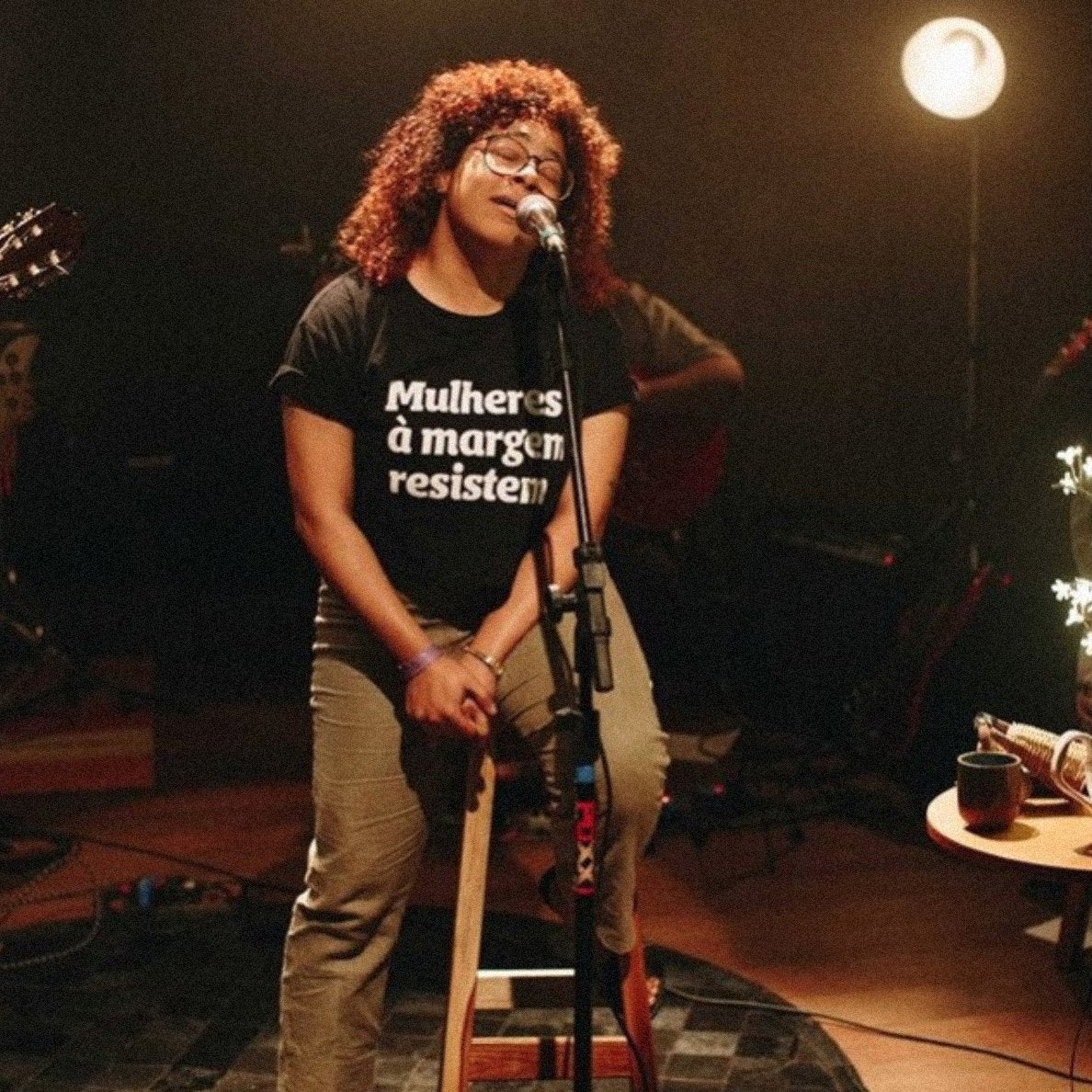 Foto colorida de uma mulher negra, cabelos castanhos, cacheados, soltos, até os ombros, usa óculos de grau, está vestida com camiseta preta escrita mulheres à margem resistem em branco. Está sentada em um banco alto, cantando no microfone.