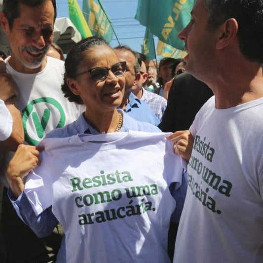 Foto colorida da Marina Silva, mulher negra, cabelos castanhos, presos em coque, com óculos de grau, segurando camiseta branca escrito resista como uma araucária em verde. Está ao lado de homem branco, cabelos curtos, castanhos, vestido com camiseta branca escrito resista como uma araucária em verde.