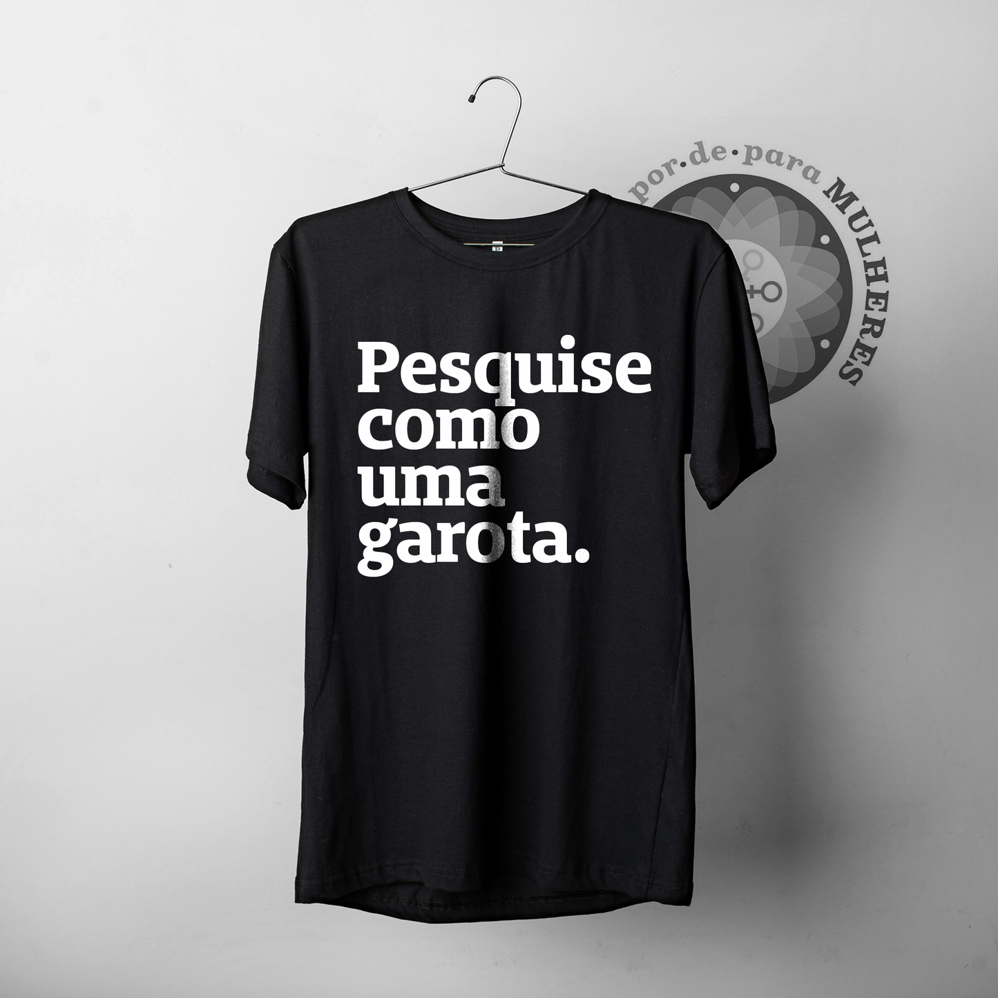 Foto preta e branca de uma camiseta preta escrita pesquise como uma garota em branco, pendurada em um cabide. Fundo branco com logo Política por de para mulheres.