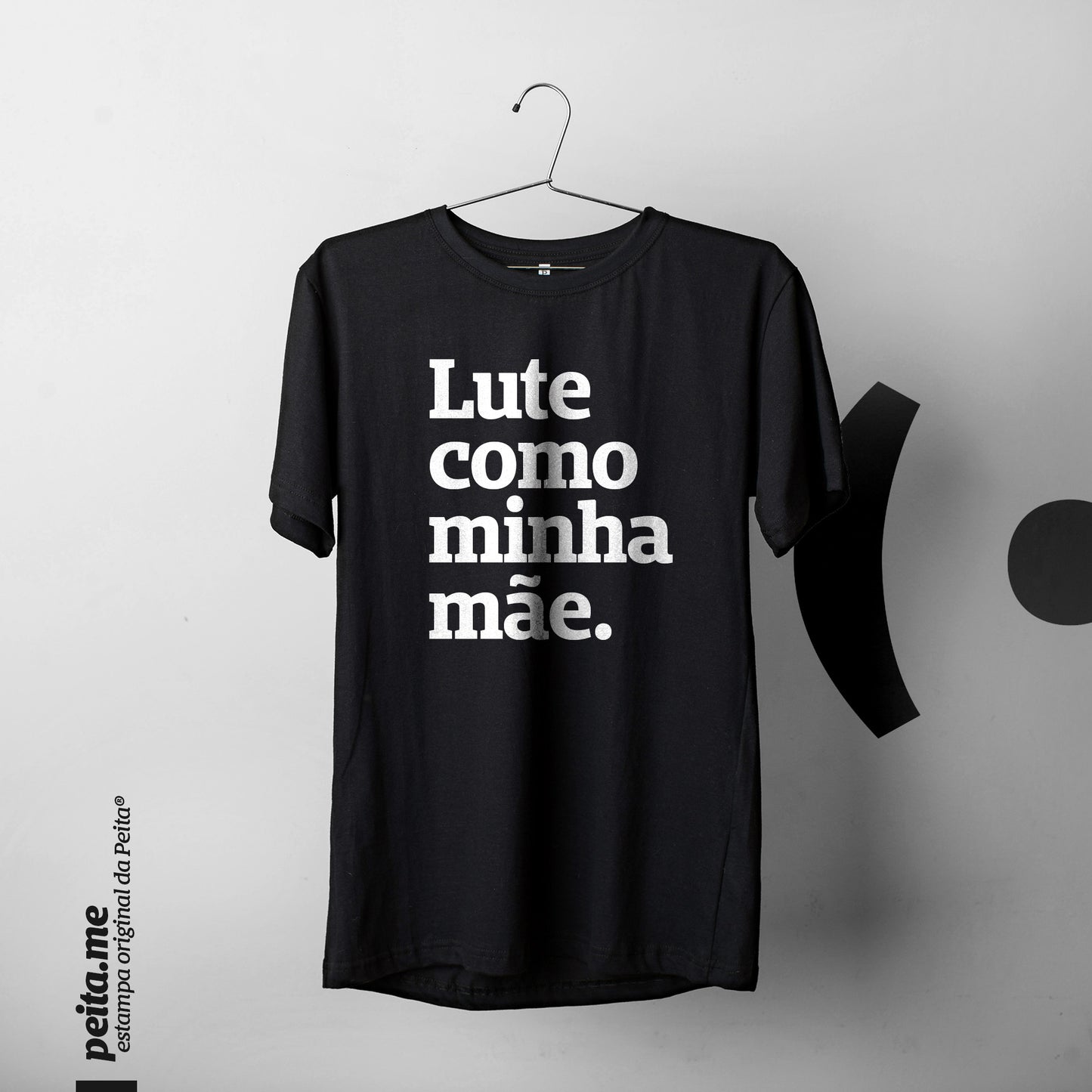  Foto colorida de uma camiseta preta escrito lute como minha mãe, pendurada em um cabide. Fundo cinza claro.