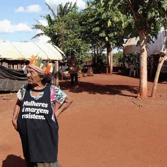 Foto colorida de uma mulher indígena, cabelos grisalhos, soltos, lisos e está vestida com camiseta preta escrita mulheres à margem resistes em branco. Está com cocar na cabeça.