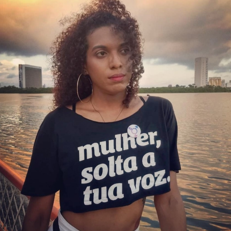 Foto colorida de uma mulher negra com cabelos castanhos, cacheados, soltos, usa brincos de argola prateado, está vestida com camiseta preta escrita mulher, solta a tua voz em branco. olha para o horizonte.