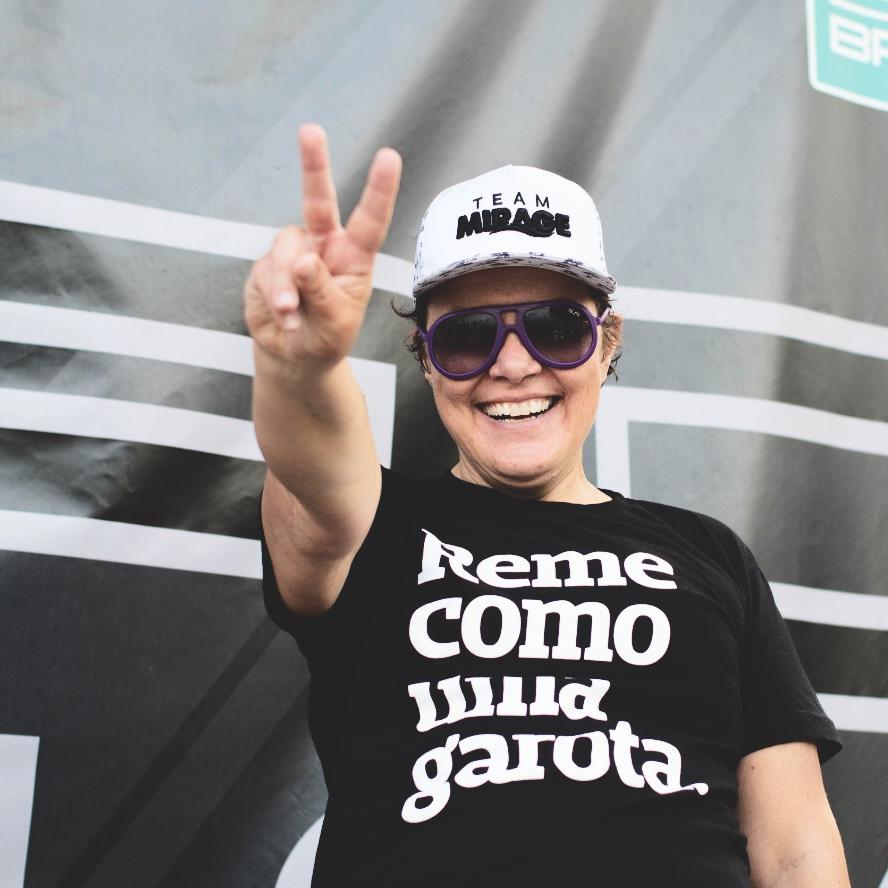 Foto colorida de uma mulher branca, cabelos curtos, castanhos, com boné branco, óculos escuro, vestida com camiseta branca escrita reme como uma garota em branco. Ela faz sinal de paz e amor com a mão esquerda, sorri na foto.