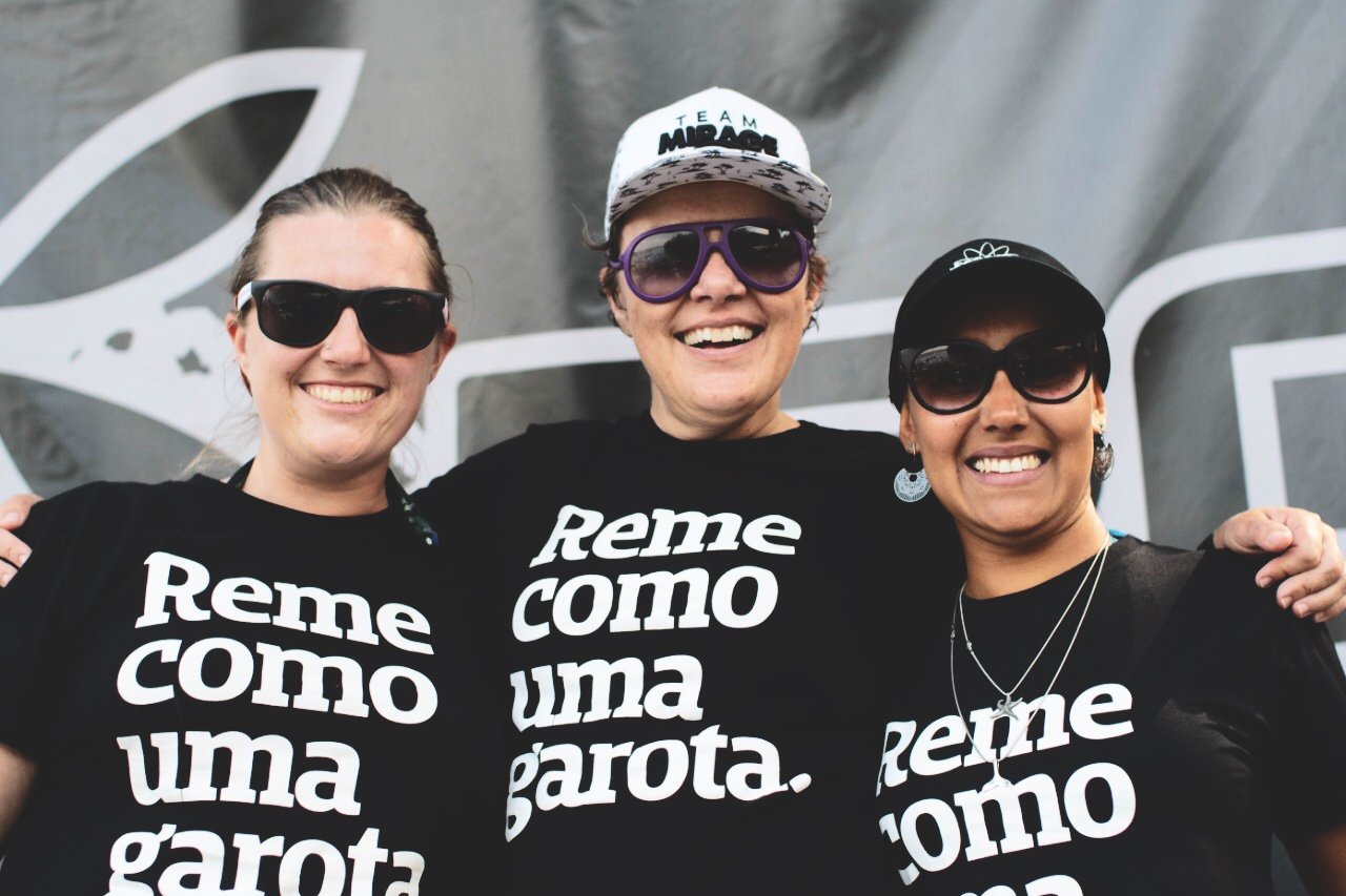 Foto colorida de 3 mulheres brancas, cabelos castanhos, presos, sendo que a do meio usa boné branco, estão vestidas com  camiseta preta escrita reme como uma garota em branco, usam óculos escuros e sorriem para foto.