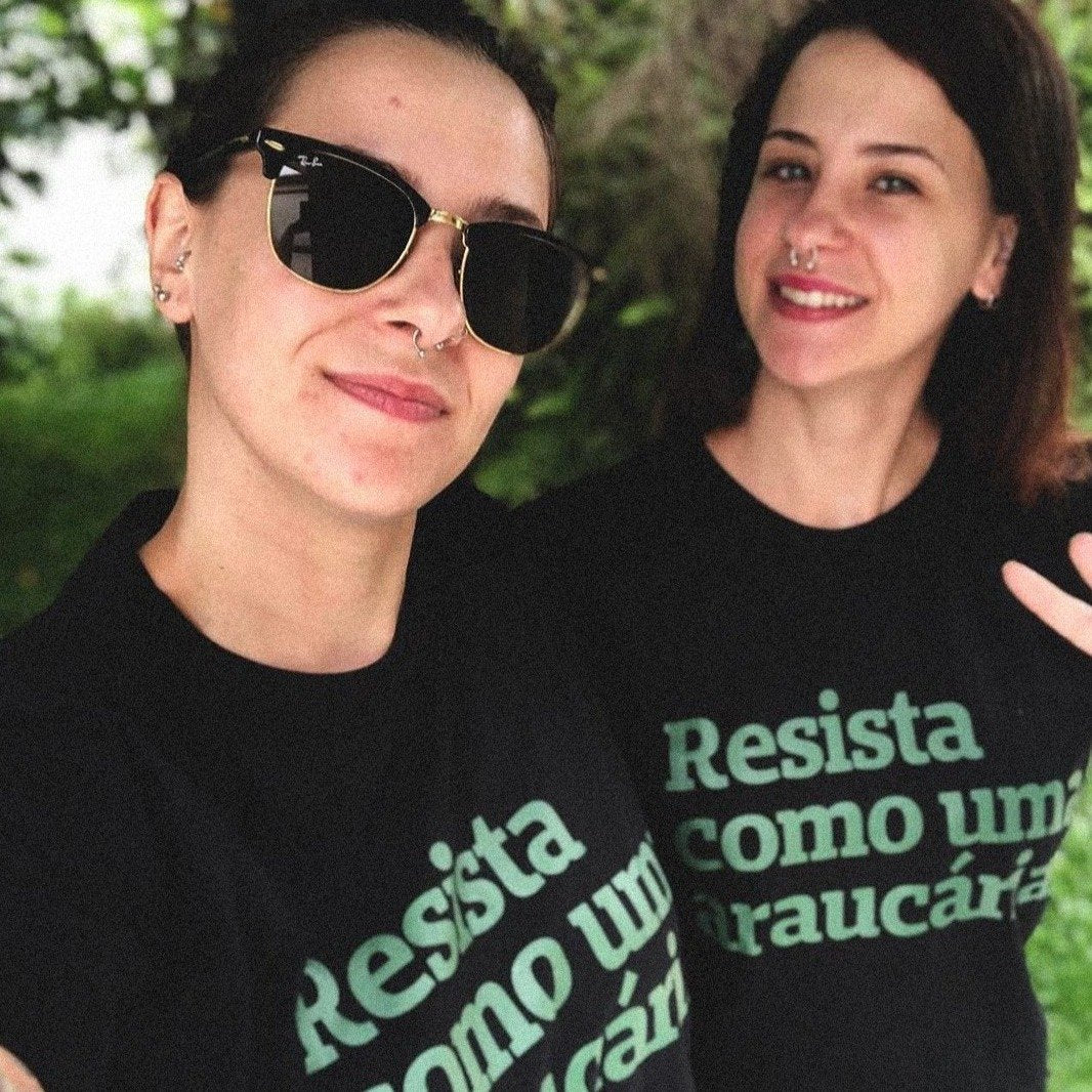 Foto colorida de 2 mulheres brancas, a da esquerda tem cabelos castanhos, lisos, presos, óculos escuros. A da esquerda tem cabelos castanhos, lisos, soltos, tem piercing no nariz. Ambas estão vestidas de camiseta preta escrita resista como uma araucária em verde. Sorriem na foto.