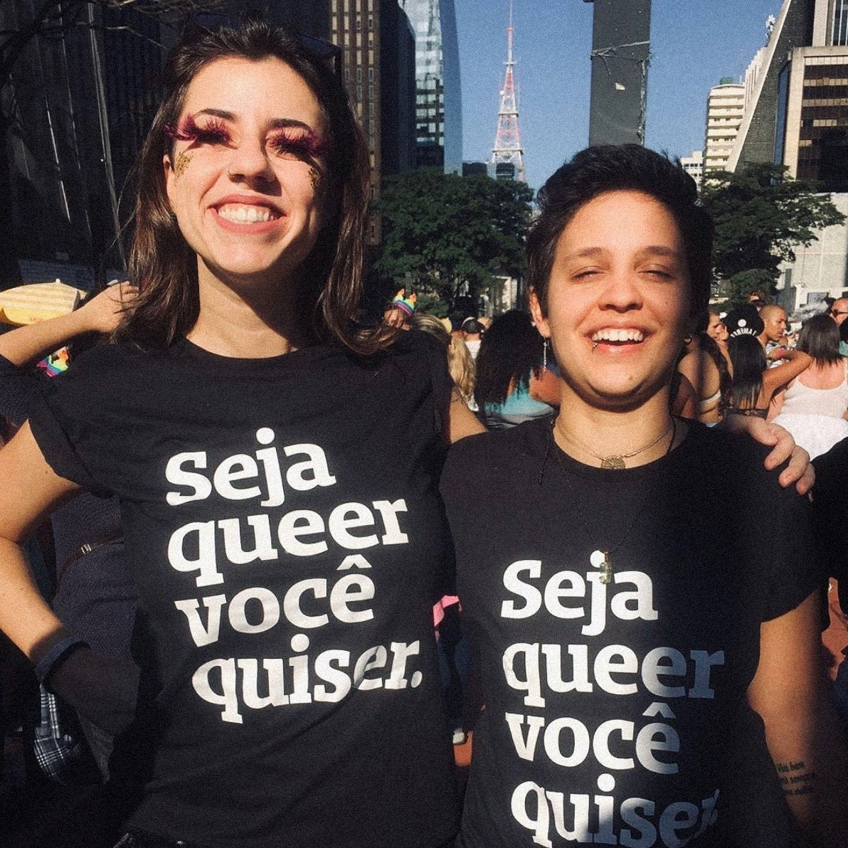 Foto colorida 2 mulheres brancas, a da direita tem cabelos castanhos, lisos, médios, está com cílios postiços de pena rosa, a da esquerda tem cabelos castanhos, curtos, lisos e tem tatuagem no braço esquerdo. Ambas estão vestidas com camisa preta escrita seja queer você quiser em branco e sorriem na foto.