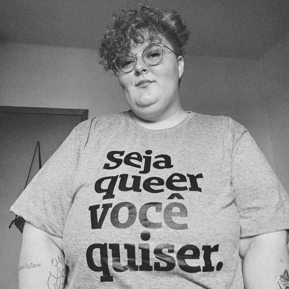 Foto preta e branca de mulher branca, cabelos claros, cacheados, curtos, usa óculos de grau, tem tatuagens nos braços e está vestida com camiseta cinza escrita seja queer você quiser em preto.