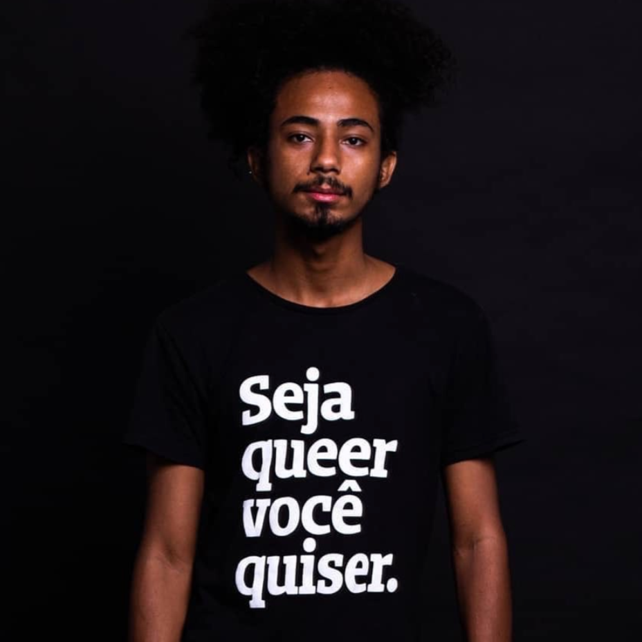 Foto colorida de homem negro, cabelo crespos, médios, presos em coque e cavanhaque, vestido com camiseta preta escrita seja queer você quiser em branco. Olha sério na foto.