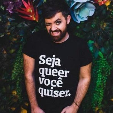 Foto colorida de um homem branco, cabelos castanhos, lisos, curtos, com barba curta, make preta nos olhos, vestido com camiseta preta escrita seja queer você quiser em branco. Ele está piscando o olho direito encostado em uma parede de flores.