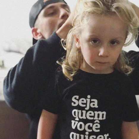 Foto colorida de um menino branco, cabelos loiros, lisos, com franja, chanel, vestida com camiseta preta escrita seja quem você quiser em branco. Está séria na foto enquanto seu pai, um homem branco com boné preto virado para trás, arruma seu cabelo.