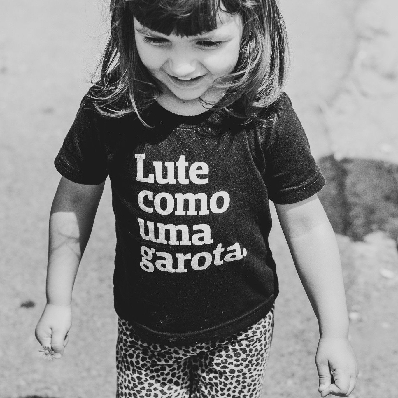 Foto preta e branca de uma menina branca, cabelos castanhos, lisos, chanel, com franja, vestida com camiseta preta escrita lute como uma garota em branco. Ela anda e sorri na foto.