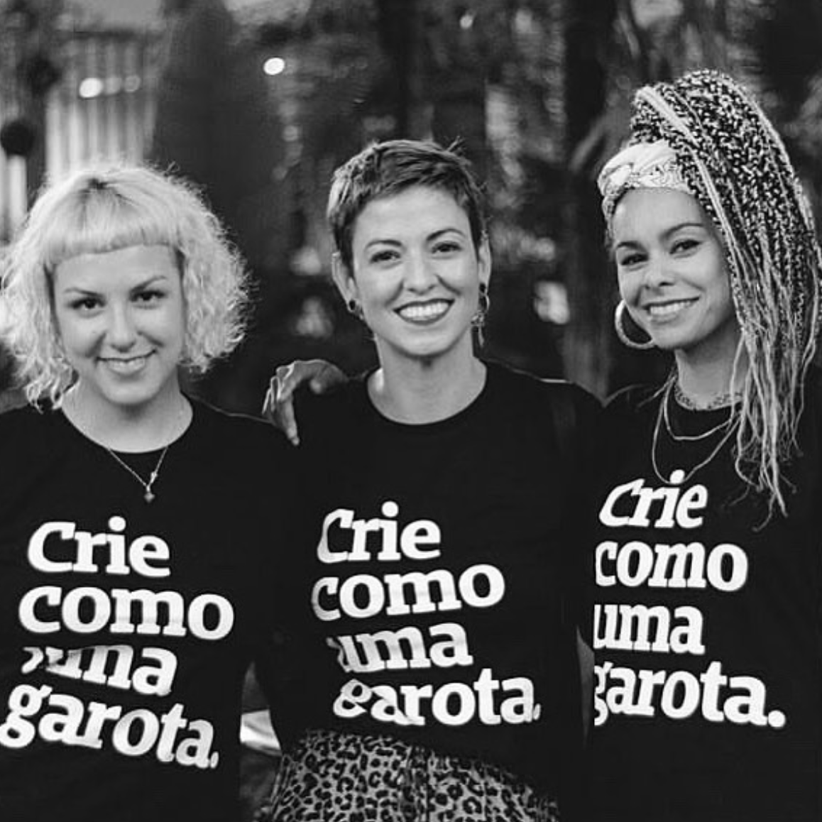 Foto preta e branca com 3 mulheres. Da direita para a esquerda: mulher negra com tranças escuras e claras no cabelo, presas em um rabo. No meio, mulher branca com cabelos curtos, lisos e escuros e a da esquerda, mulher branca com cabelo chanel, platinado. Todas estão sorrindo na foto e abraçadas. Elas vestem peita preta com a frase crie como uma garota em branco.  Fundo desfocado.