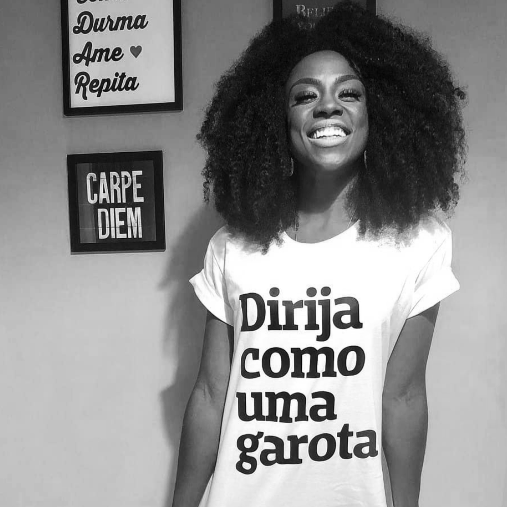 Foto preta e branca de uma mulher negra, cabelos crespos, soltos, médios e castanhos. Vestida com camiseta branca escrita dirija como uma garota em preto. Ela está sorrindo, puxando a barra, encostada em uma parede com quadros pendurados.