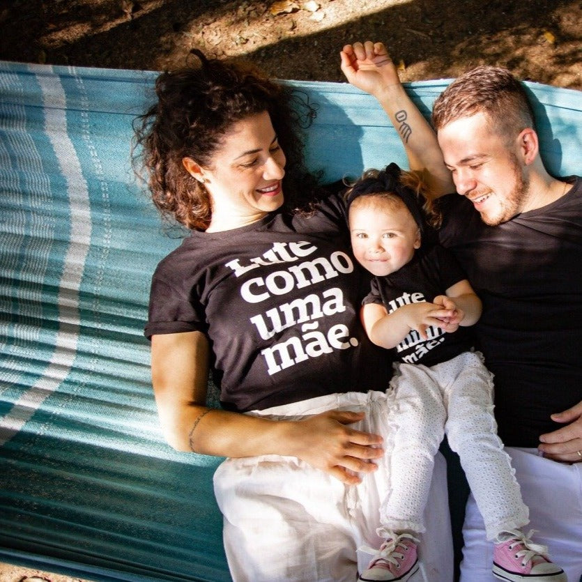 Foto colorida de uma família deitados em uma rede, a mãe mulher branca de cabelos cacheados escuros veste uma camiseta rpeta com frase Lute como uma mão em branco, a filha no meio do casal criança branca  de cabelos claros veste peitinha preta com frase Lute como minha mãe em brando, usa faixa preta na cabeça e morde lábio inferior e sorri. Pai homem branco de cabelos curtos e claros, veste camiseta preta lisa. O casal olha e sorri para a criança no meio. todos de vestem calça branca. 